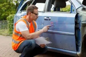 A man in an orange safety vest inspects and documents damage on the side of a blue car with a clipboard and pen.