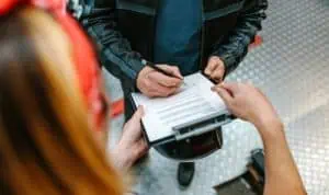 Person filling out or signing a document on a clipboard held by another individual, both standing indoors on a patterned metal floor.