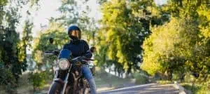 A person wearing a helmet and dark clothing sits on a motorcycle parked on a road lined with trees in daylight.