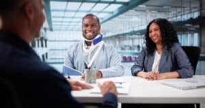 A man with a neck brace and arm in a sling sits at a desk with a woman, speaking to another person in an office setting.