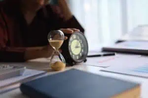 A person sits at a desk with an hourglass and an analog clock, surrounded by papers and a closed book.